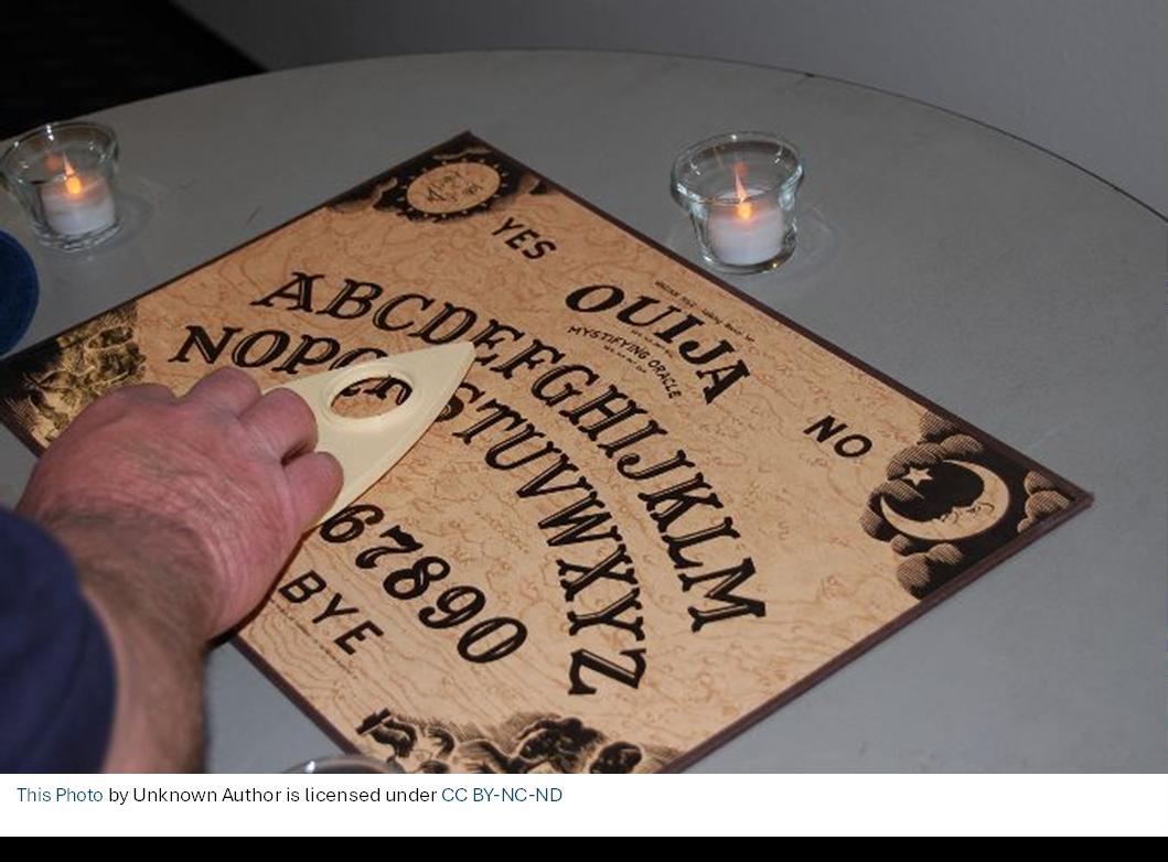 Hand using a planchette on a Ouija board with candles on the table.