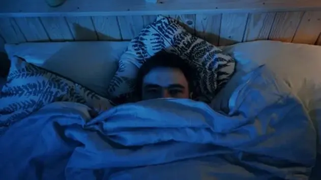 Man lying in bed under covers with wide eyes, staring fearfully in dim blue light—evoking a scared reaction at 3AM.