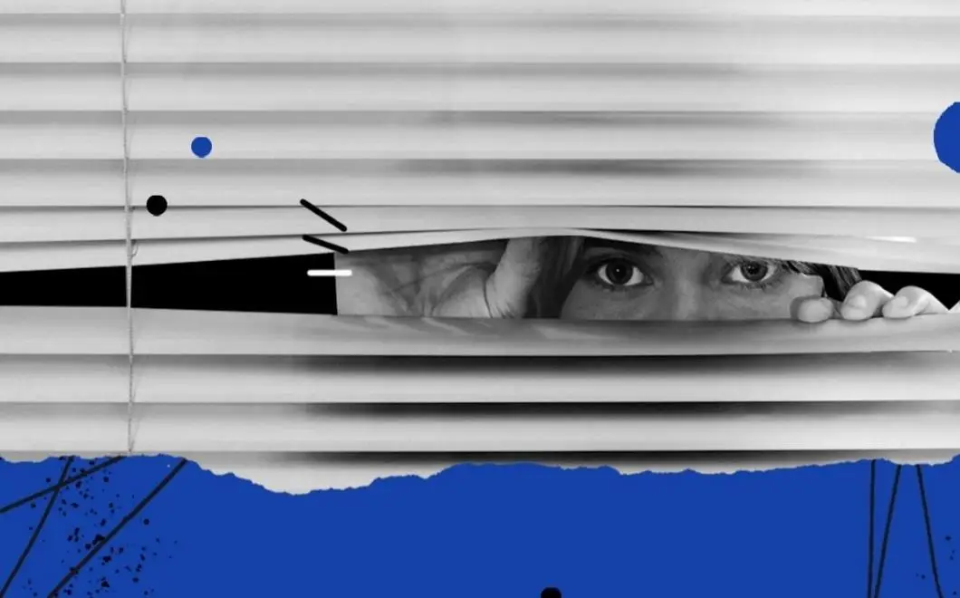 Person peering through window blinds at night, illustrating one of the common signs your house is haunted – feeling watched.